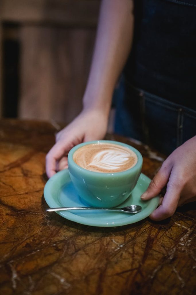 pexels photo 13737060 Close-up of a woman holding a blue cup with latte art on a wooden table.