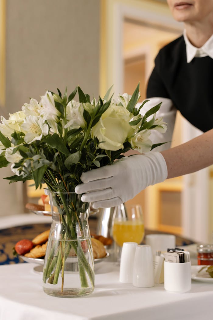 services-04 Woman arranging flowers in a hotel room with breakfast on a table, emphasizing luxury and convenience.